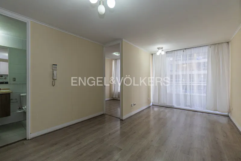 Empty living room with light beige walls, wood-look flooring, a doorway to a bathroom, and large windows with white curtains.
