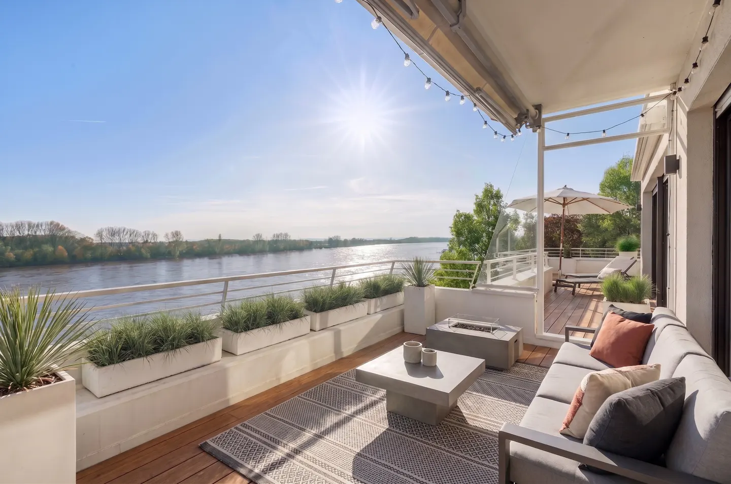 Bright, sunny patio with river view. Gray sofa, fire pit, and planters decorate the wood deck. String lights hang above.