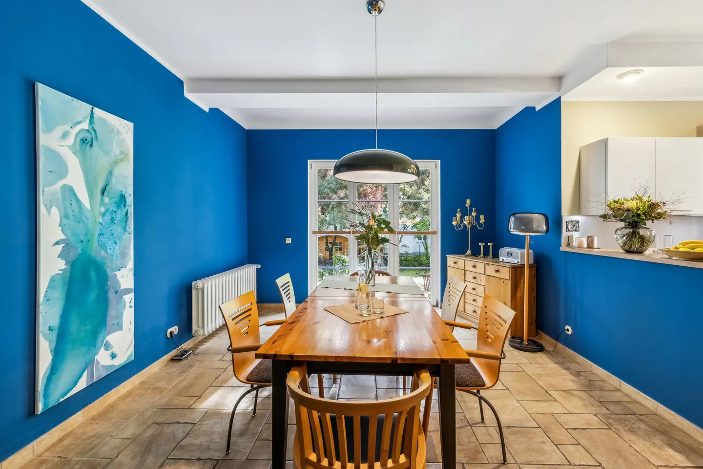 Bright blue dining room with a wood table, chairs, and abstract art. A large window lets in natural light.