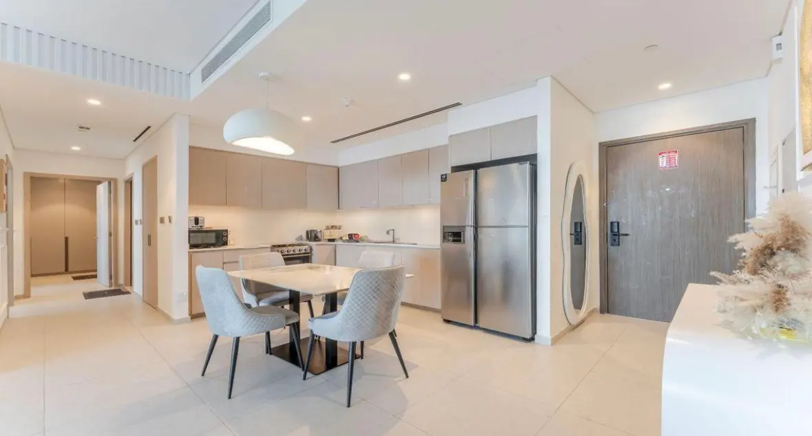 A bright, modern apartment interior with a dining table, kitchen, stainless steel refrigerator, and hallway.