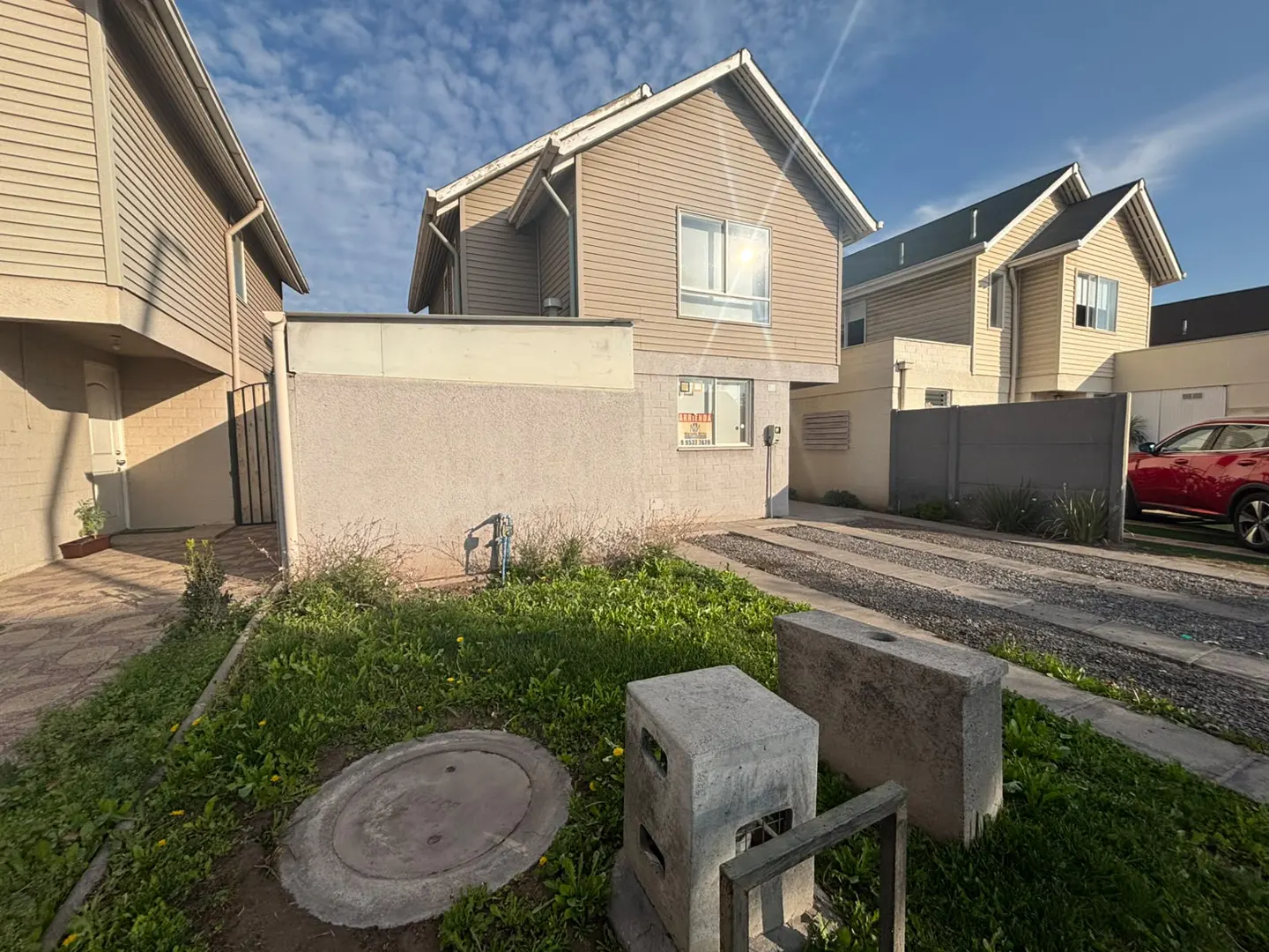 Exterior view of a beige two-story house with a gravel driveway, small front yard, and a red car parked on the right.