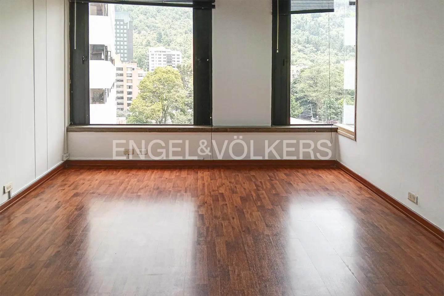 Empty room with hardwood floors, white walls, and three large windows with a city view. "Engel & Völkers" is printed on the wall.