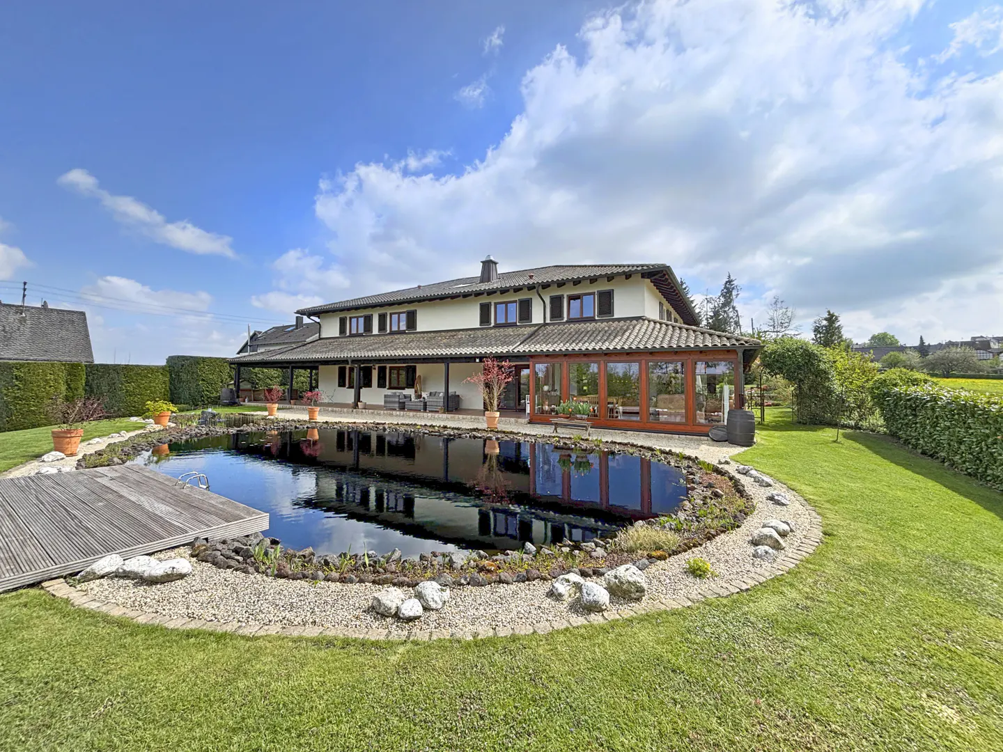 A large, cream-colored house with a pond in the backyard on a sunny day. The pond is surrounded by rocks and grass.