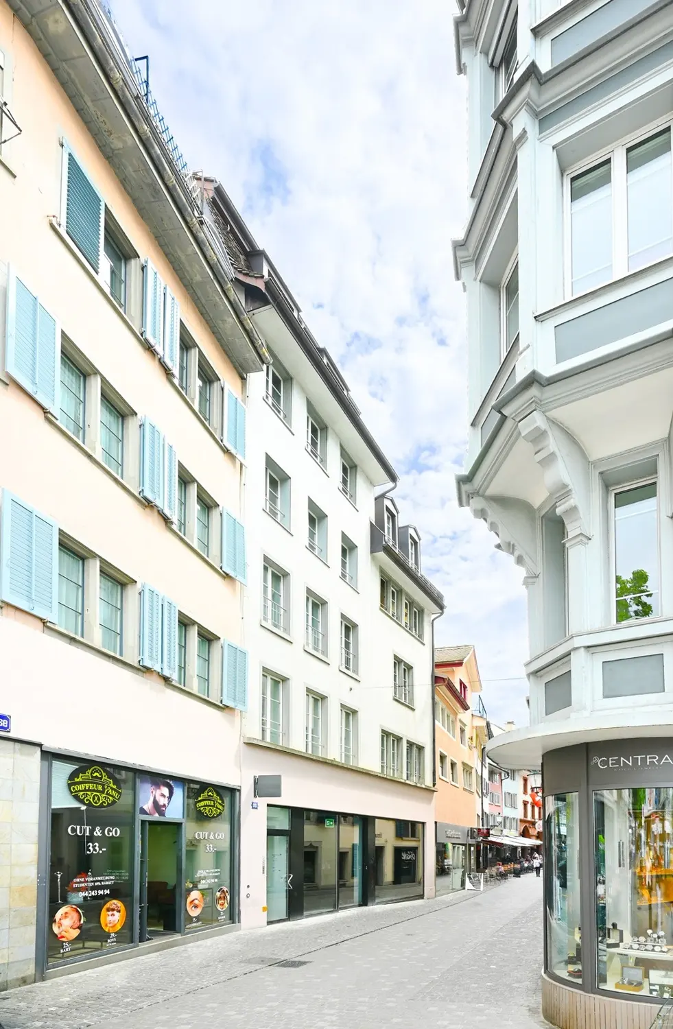 European street view with light-colored buildings, some with blue shutters, and storefronts at street level.