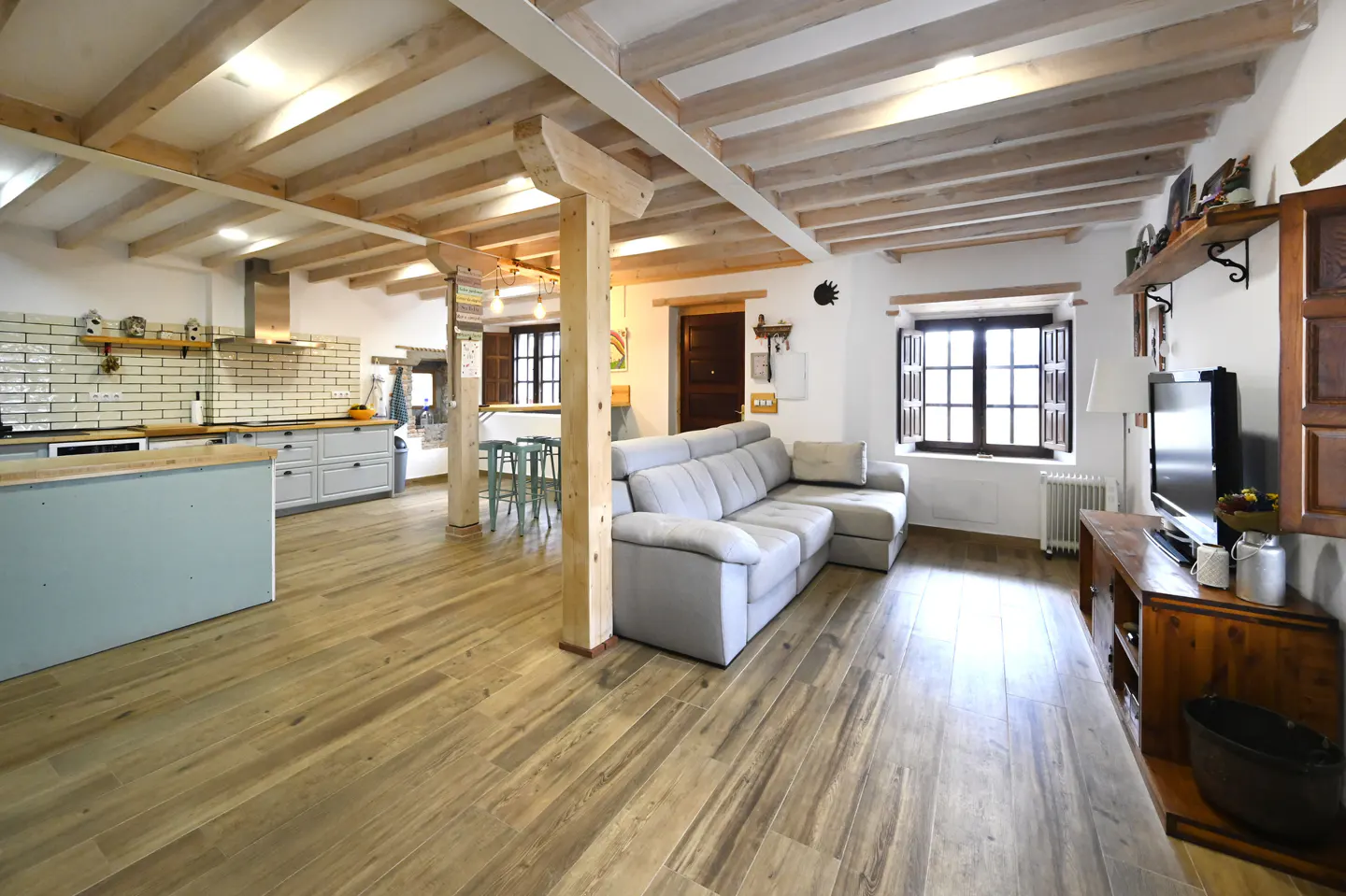 Open-concept living space with wood floors, exposed beams, and a gray sectional sofa. Kitchen island and bar stools are visible.