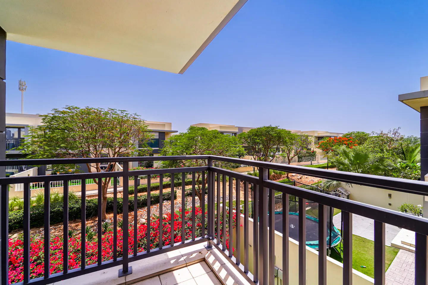Balcony view with black metal railings overlooking a garden with red flowers, green trees, and houses under a clear blue sky.