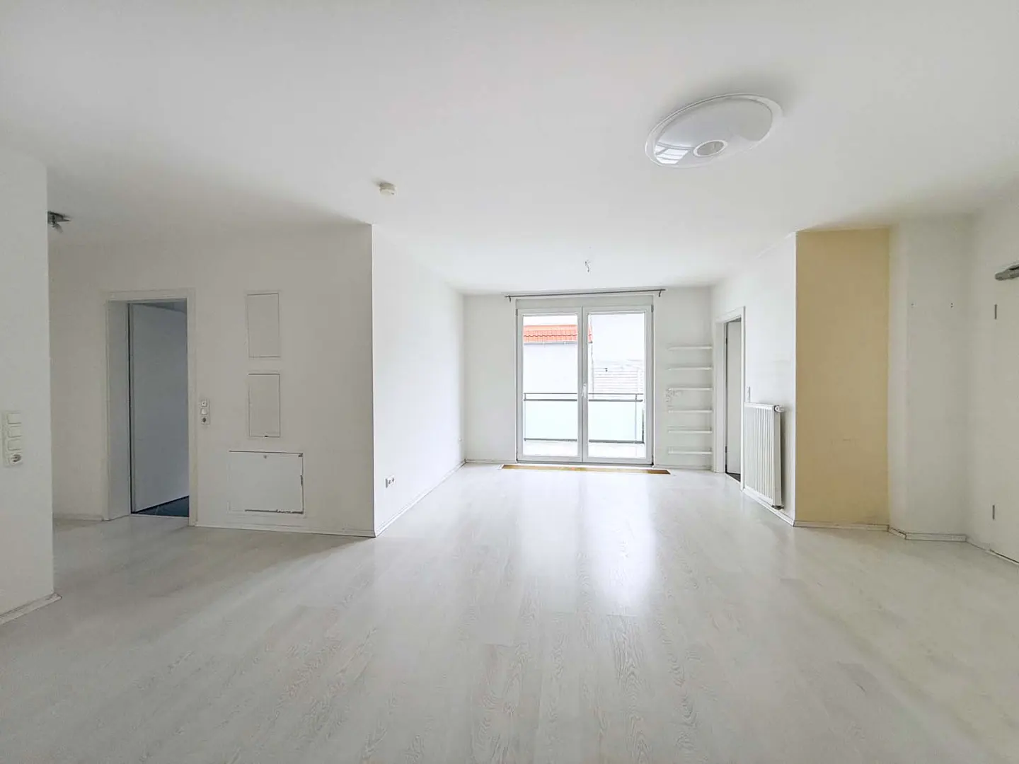 Bright, empty room with white walls, light wood floors, and a sliding glass door to a balcony. A doorway and radiator are visible.