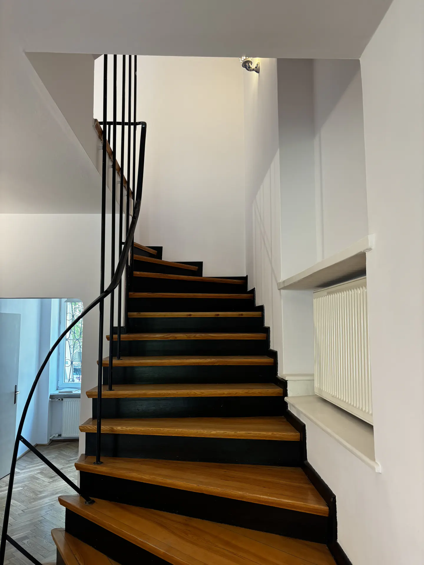 Interior view of a curved staircase with wood treads, black risers and a black metal railing in a white room.