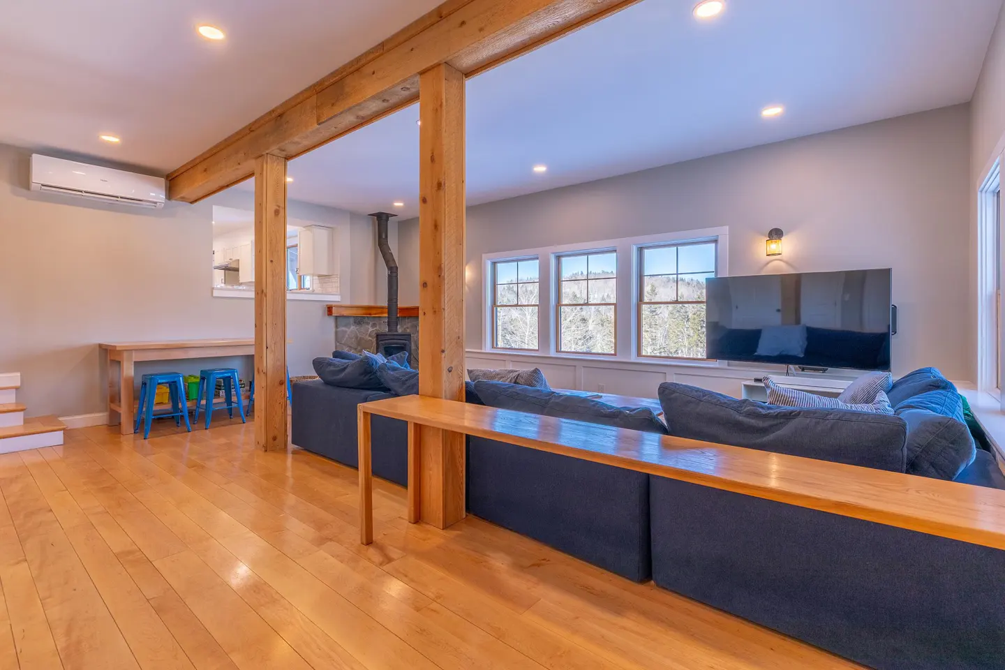 Living room with wood floors, exposed beams, and a large blue sectional sofa. Windows overlook a snowy landscape. A wood-burning stove is visible.