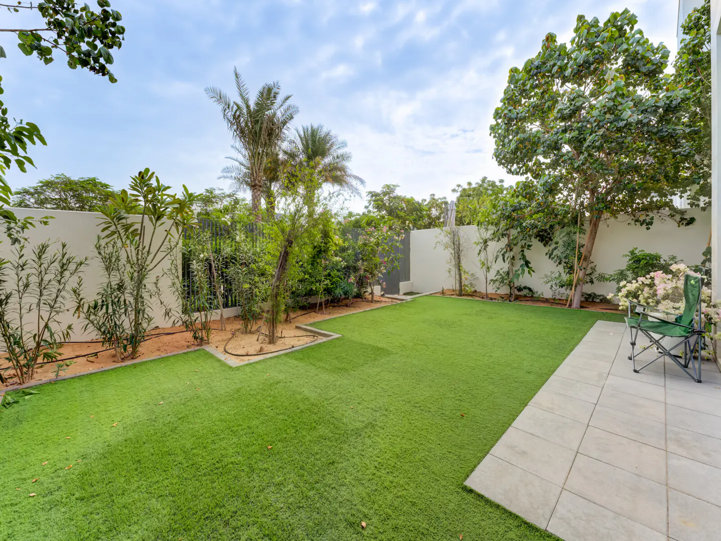 A backyard with green grass, trees, and a white wall under a cloudy sky. A green folding chair sits on a tiled patio.