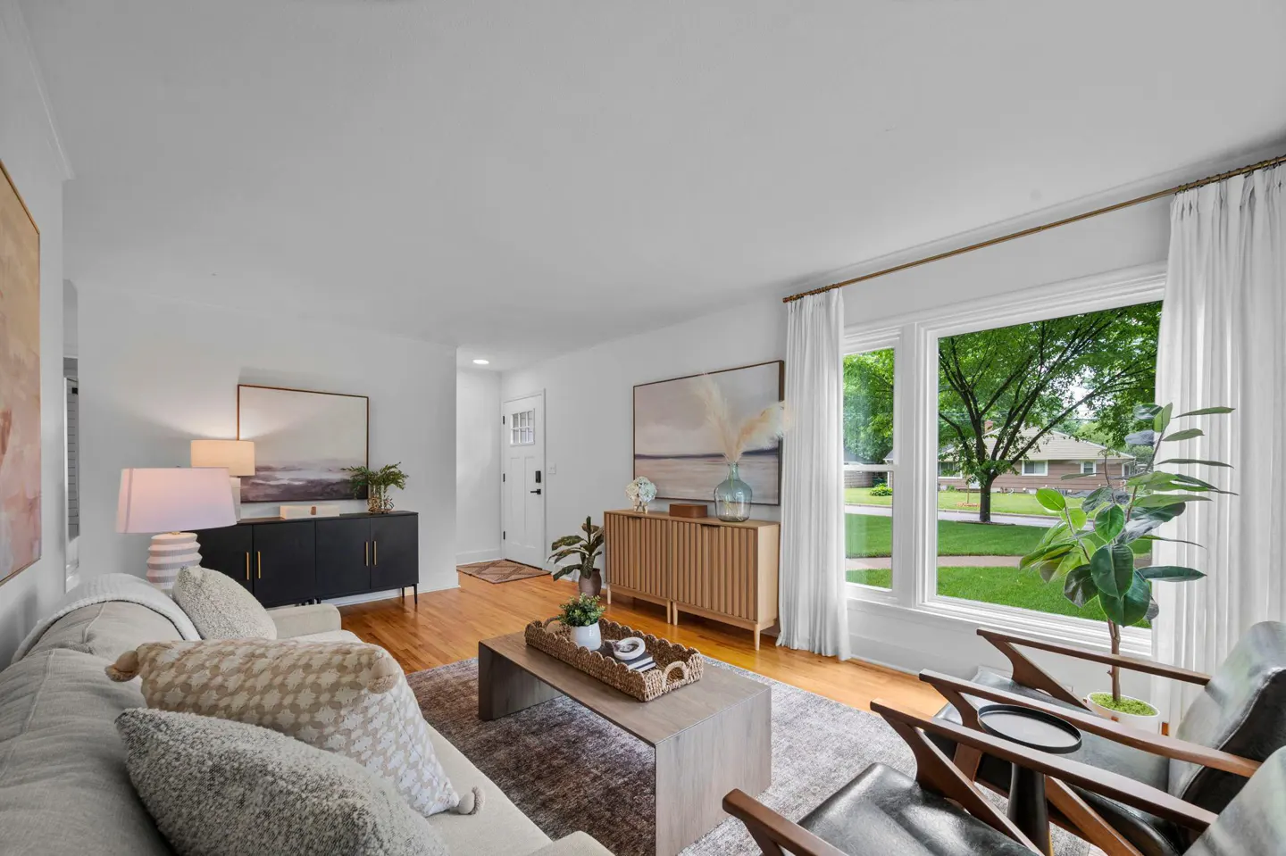 Bright living room with hardwood floors, a beige sofa, two black armchairs, a coffee table, and a large window view.