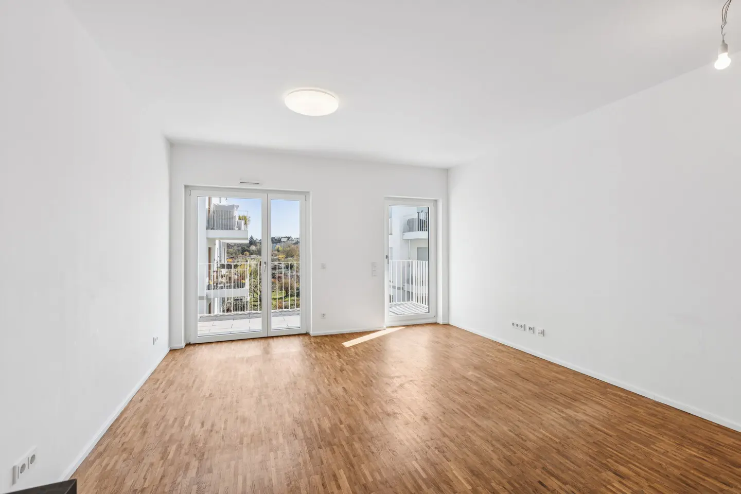 Bright, empty room with wood floors and white walls. Glass doors lead to balconies with views of trees and buildings.