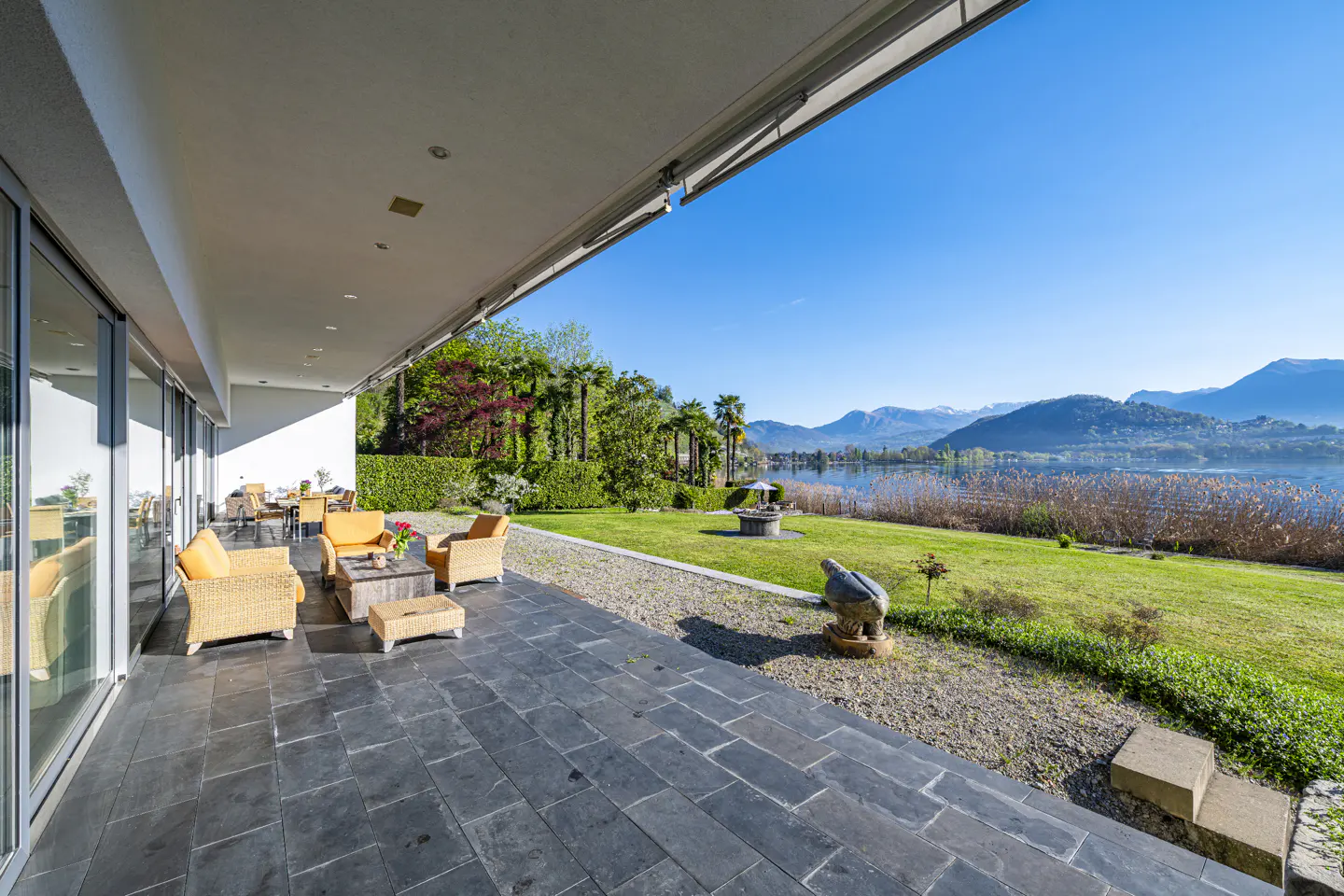 Outdoor patio with wicker furniture overlooking a lake and mountains under a blue sky.