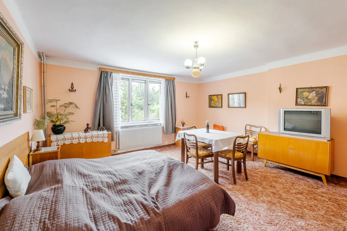 Bedroom with a brown bed, dining table with white tablecloth, and a vintage TV on a wooden cabinet. Peach walls and patterned carpet.