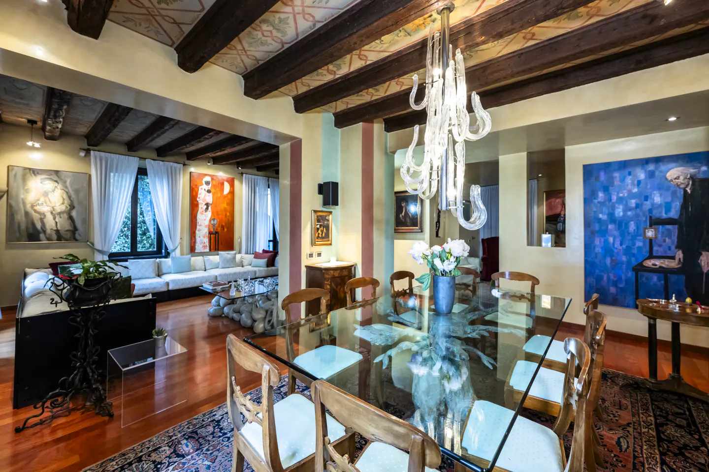 Open-concept dining and living room with wood floors, exposed beams, and colorful artwork. A glass table and chandelier are in the foreground.