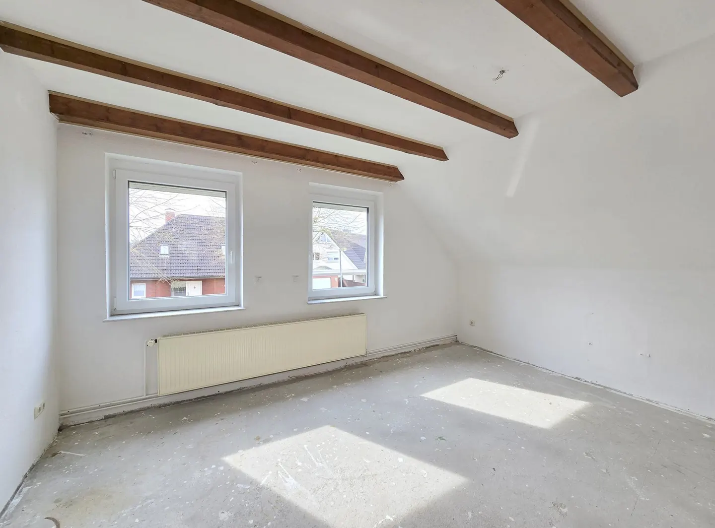 Bright, empty attic room with white walls, exposed brown beams, and two windows showing houses outside. A white radiator is under the windows.