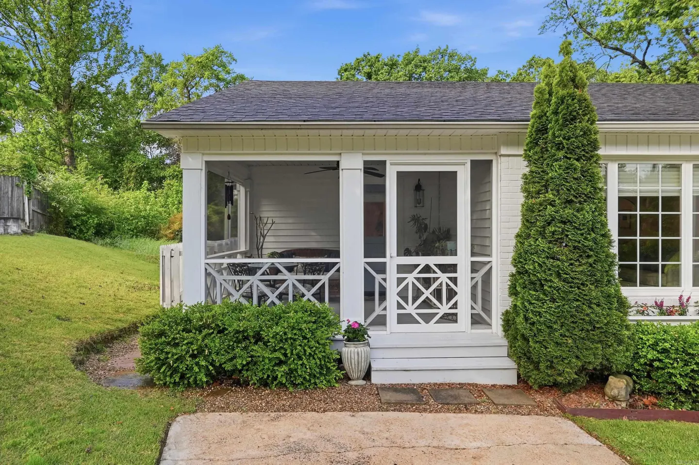 Exterior view of a white house with a screened-in porch, green shrubbery, and a tall evergreen tree.