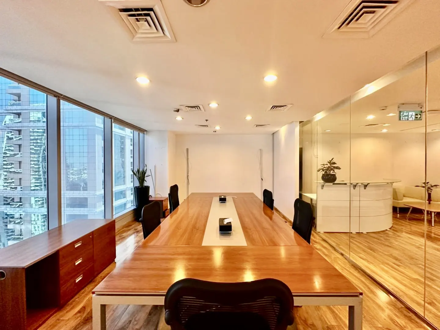 Modern office conference room with a long wooden table, black chairs, and floor-to-ceiling windows overlooking city towers.