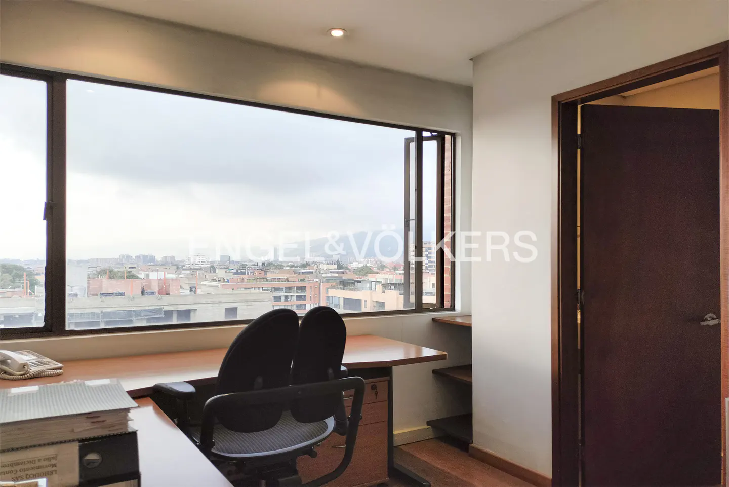 Office space with a large window overlooking a cityscape. A desk and chair are in front of the window. A closed door is on the right.