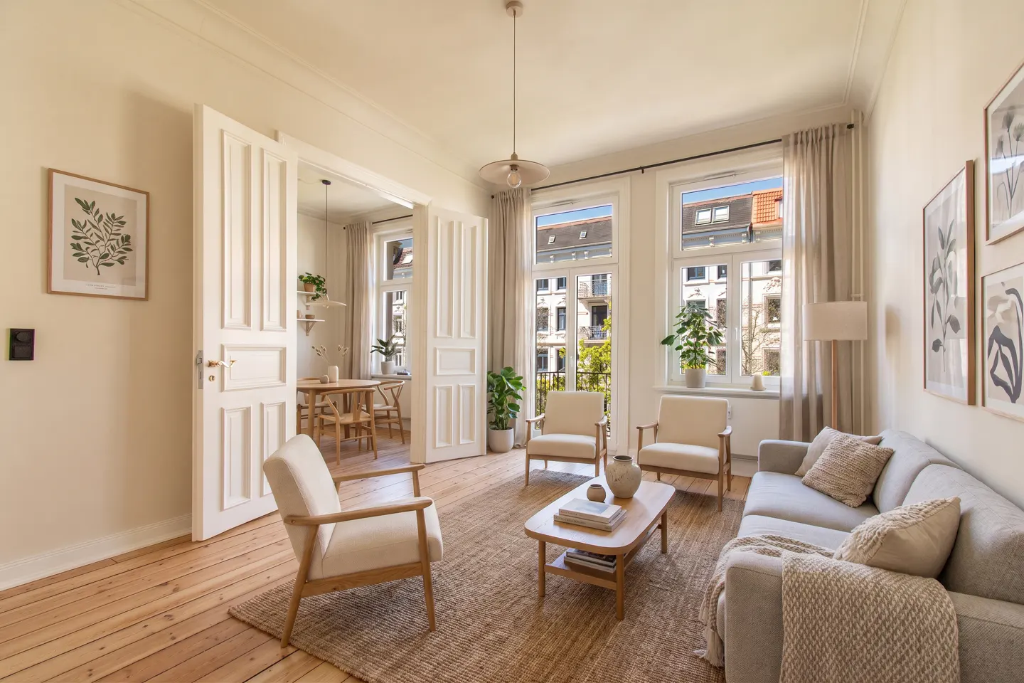 Bright living room with wood floors, a gray sofa, and cream-colored chairs. Large windows overlook a city street. Open doors lead to a dining area.