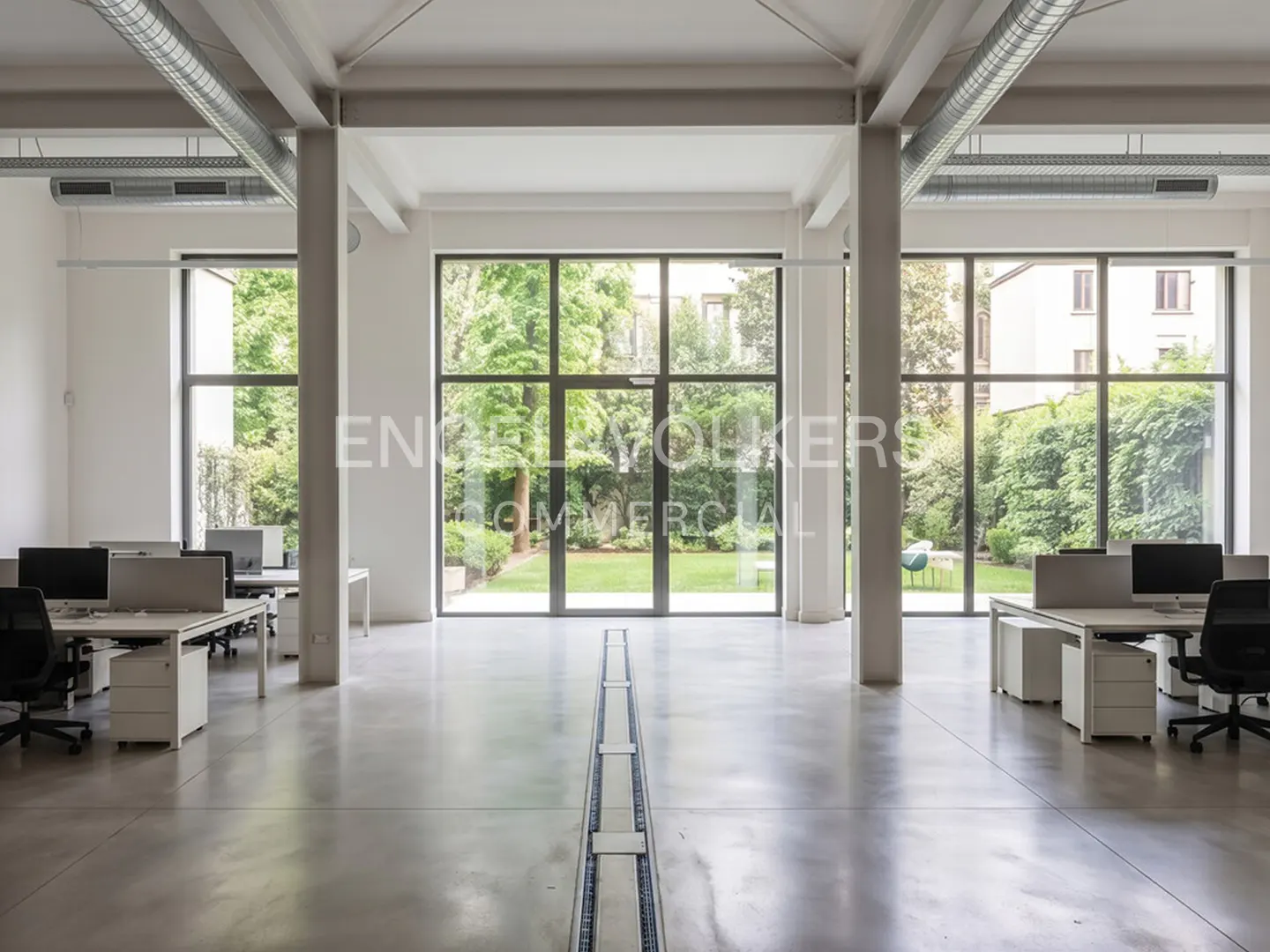 Bright, open office space with desks, computers, and large windows overlooking a green garden. White walls and concrete floors.