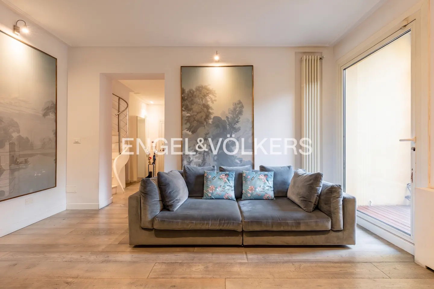Living room with a gray sofa, floral pillows, and landscape art. Light wood floors and an open doorway to a balcony.