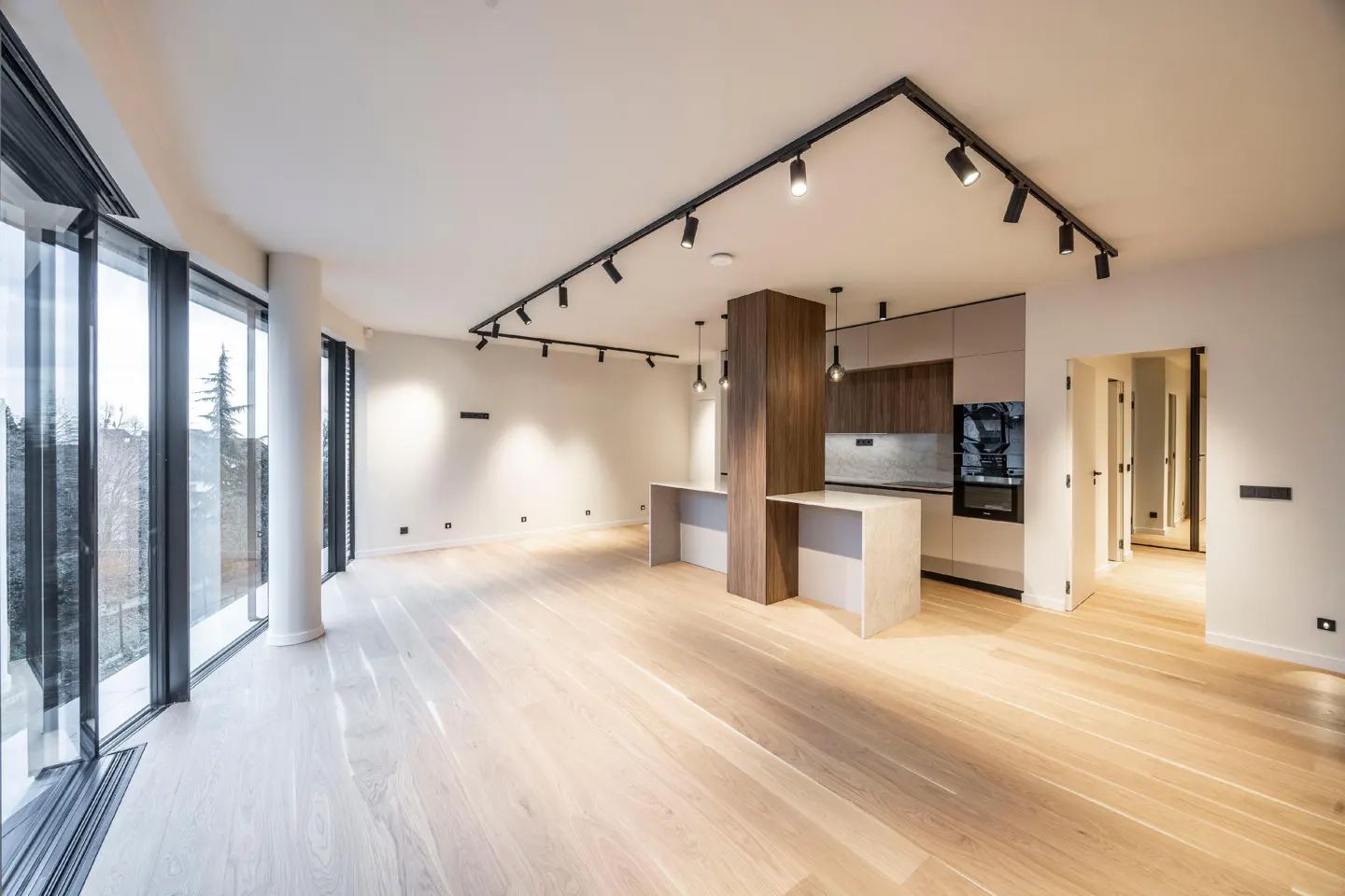 Bright, modern open-plan kitchen and living area with light wood floors, track lighting, and large windows overlooking trees.