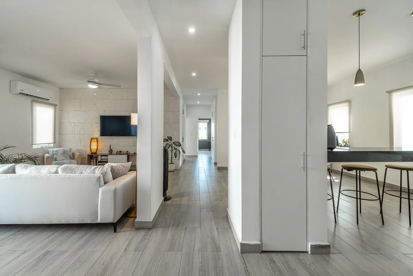 Bright, modern home interior with gray wood floors. A white sofa faces a TV. A hallway leads to a kitchen with bar stools.