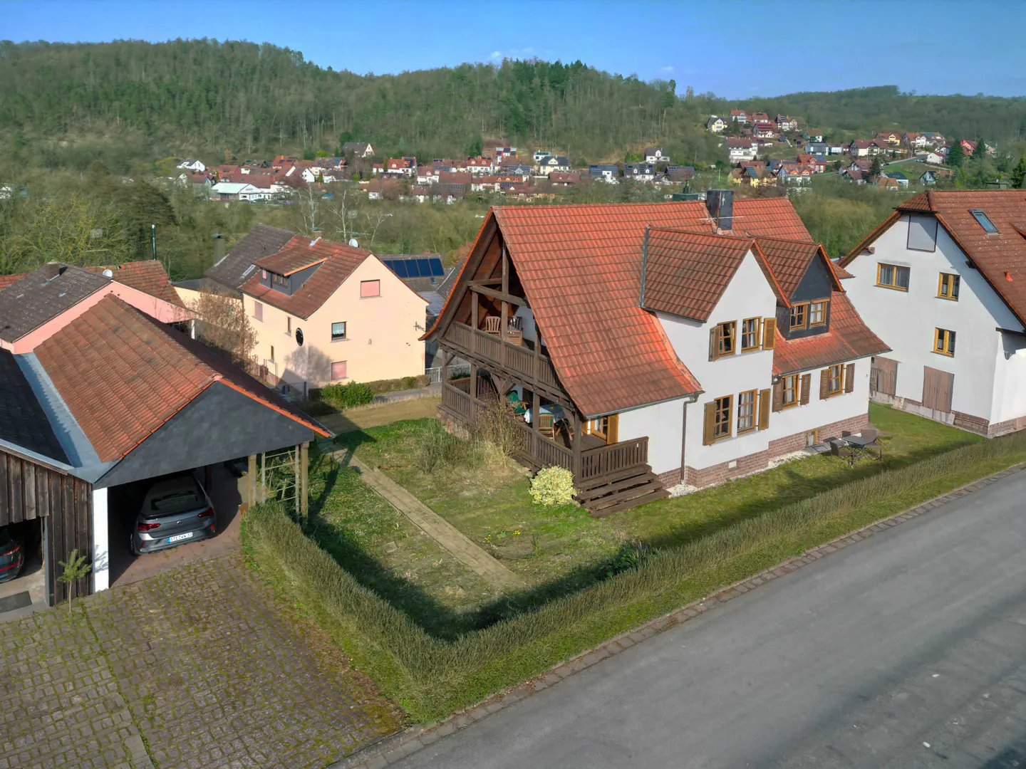 Aerial view of a white house with a red tile roof and brown shutters, surrounded by green grass and trees. A car is parked in a carport.