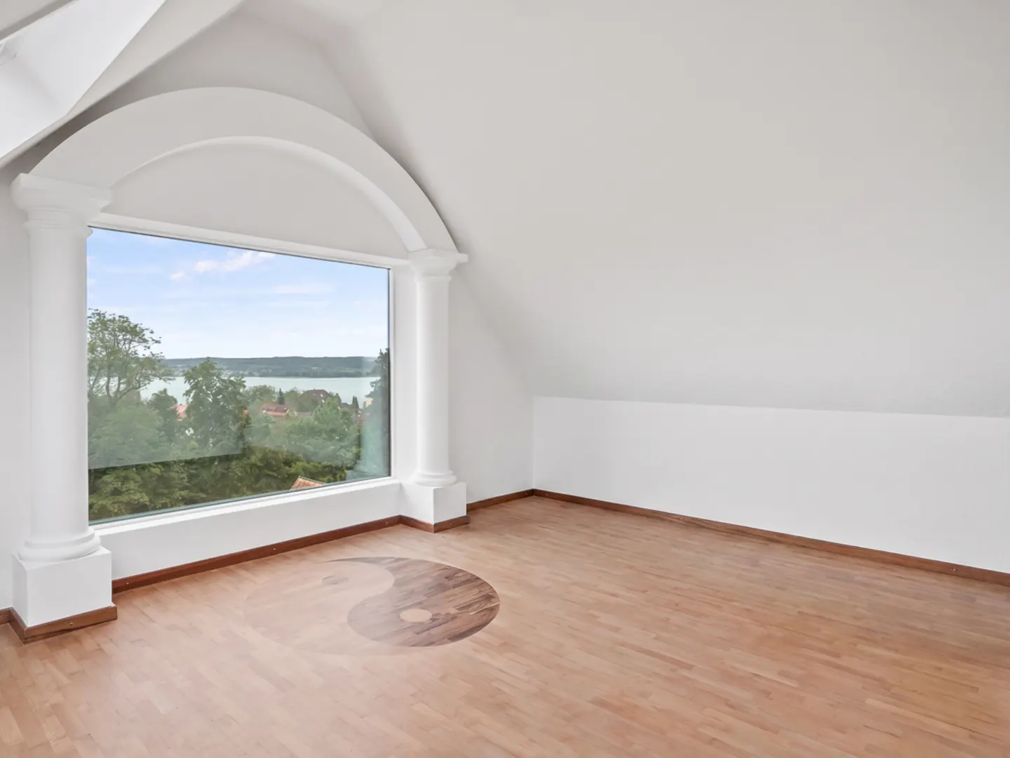 Attic room with white walls, wood floors with a yin and yang symbol, and a large arched window with a lake view.
