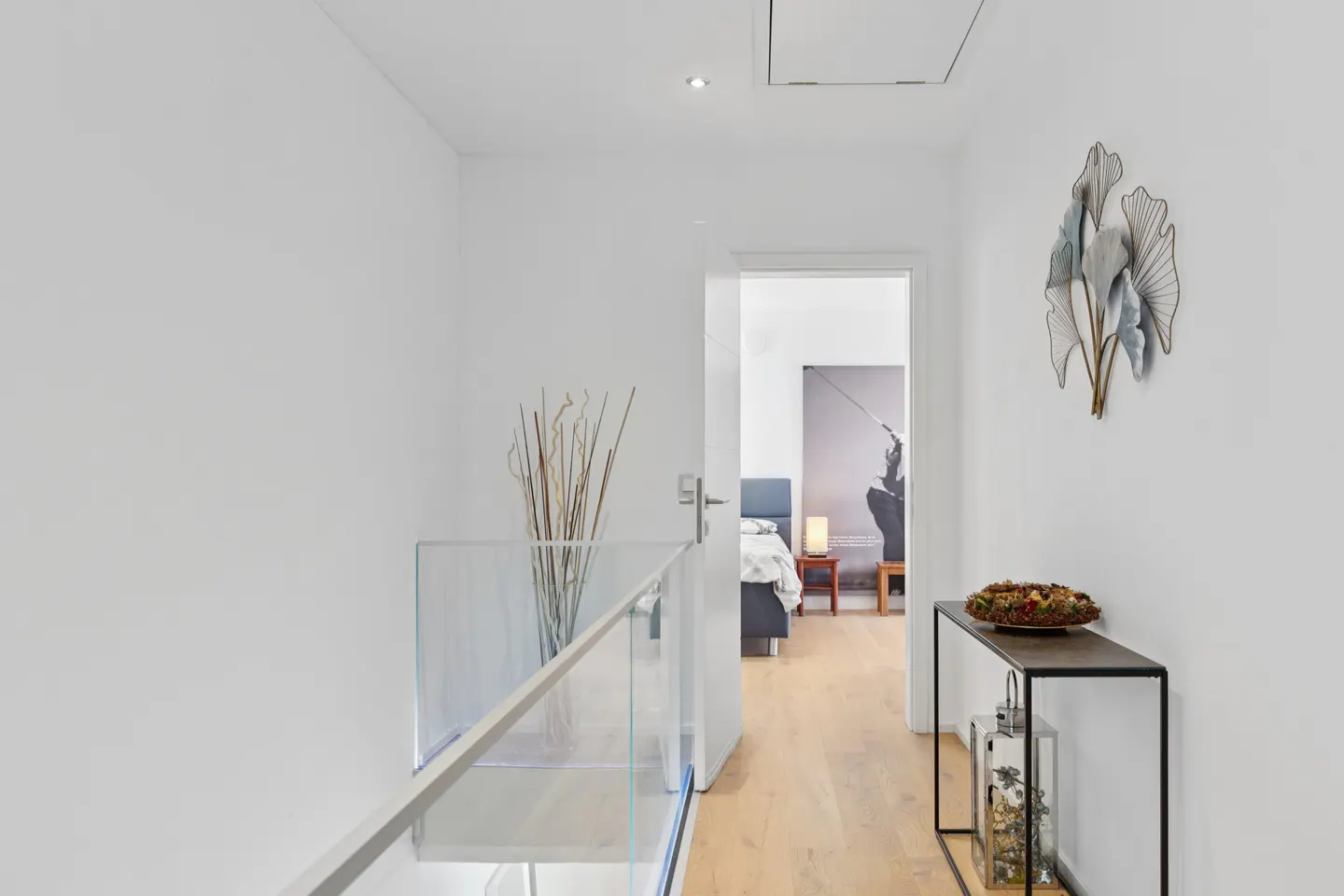 Hallway with white walls, light wood floors, and glass railing. A bedroom is visible through an open door. A console table with decor is on the right.