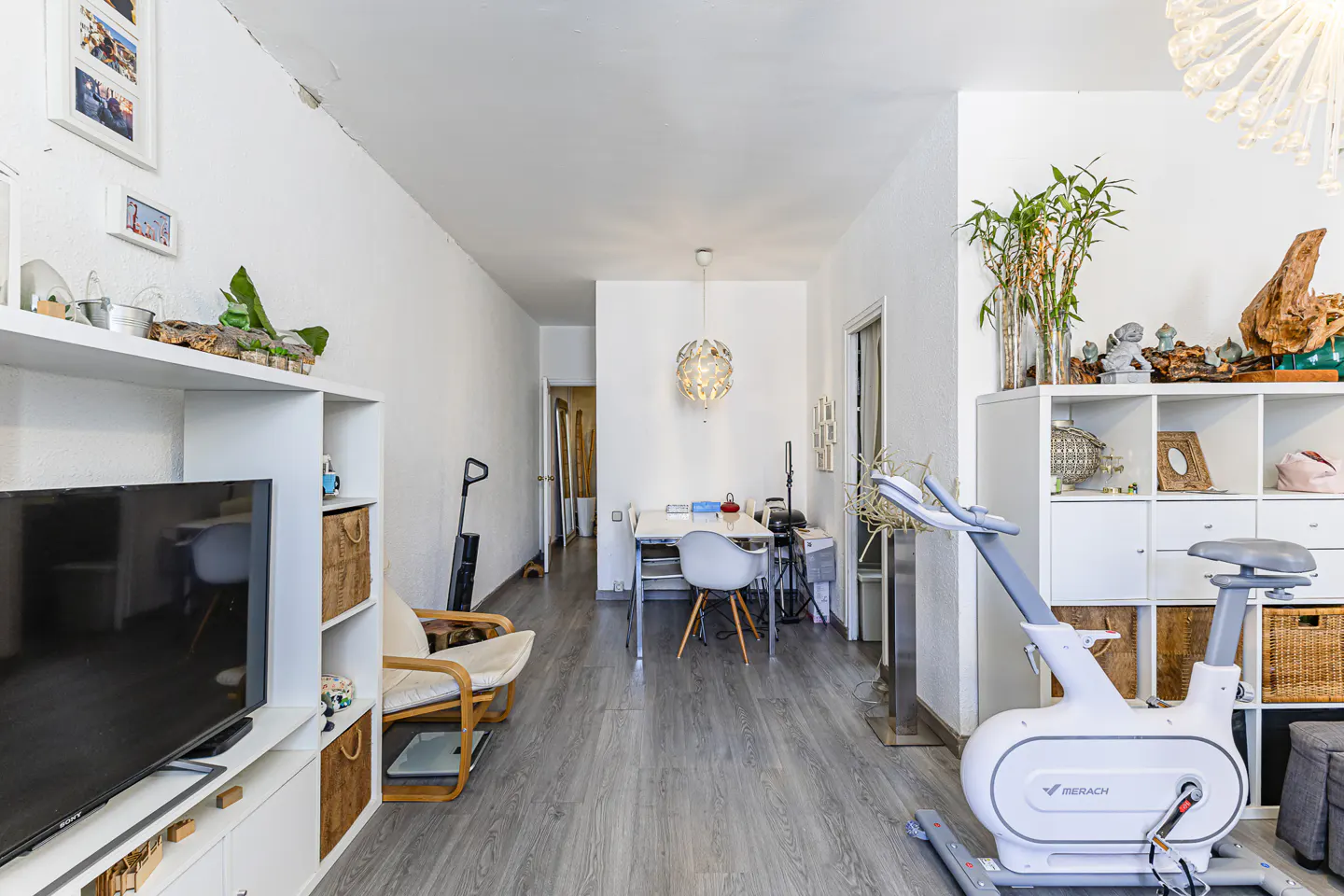 Bright living room with gray wood floors, white walls, and white shelving units. A stationary bike sits in the foreground.