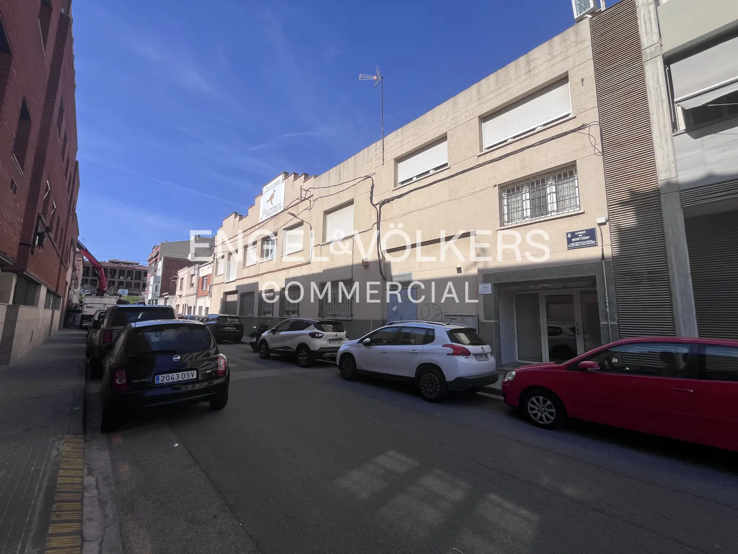 Street view of a two-story commercial building with parked cars. The building is beige with white window shades. A red car is parked in front.