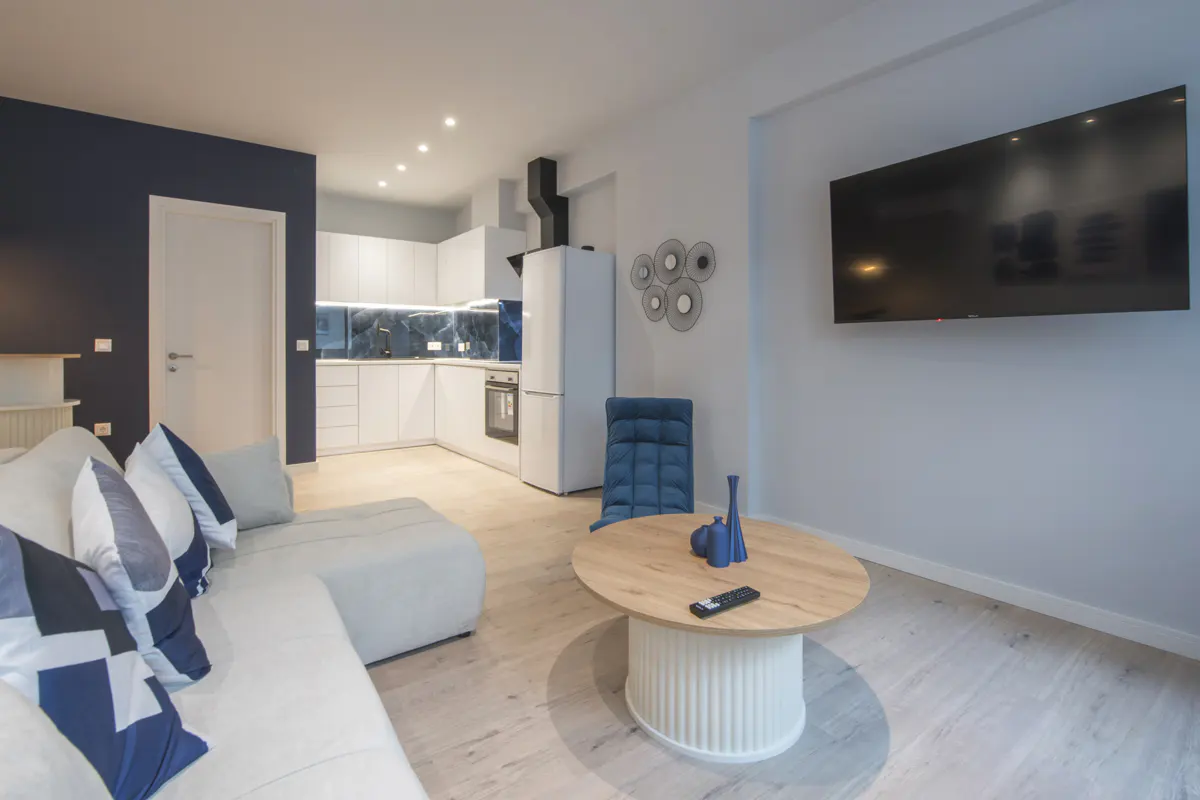 Bright, modern apartment with white sofa, round wood table, blue chair, and white kitchen in the background.