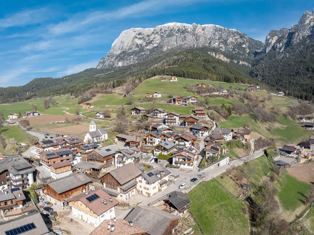 Aerial view of a village with brown-roofed houses, green hills, and a snow-capped mountain range in the background.