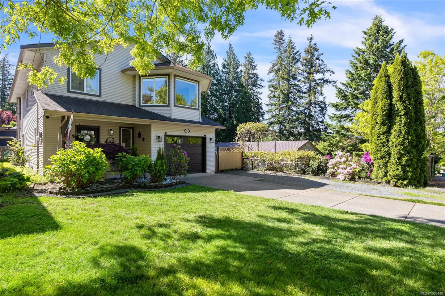 Two-story beige house with a dark roof, green lawn, and driveway leading to a garage. Trees and shrubs surround the property.