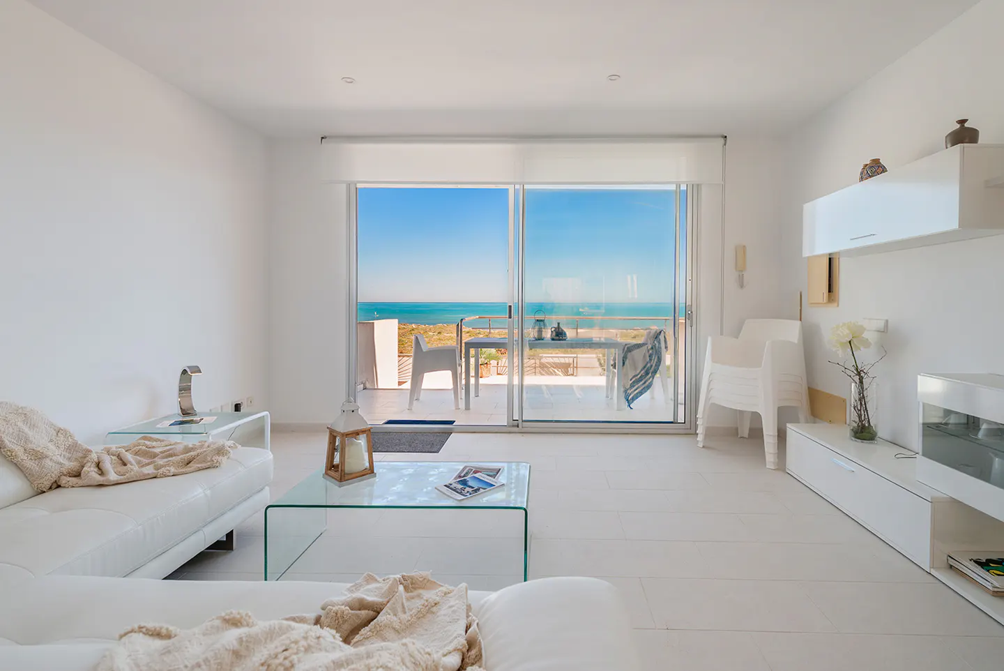 Bright, all-white living room with a glass coffee table and a view of the ocean from the balcony.