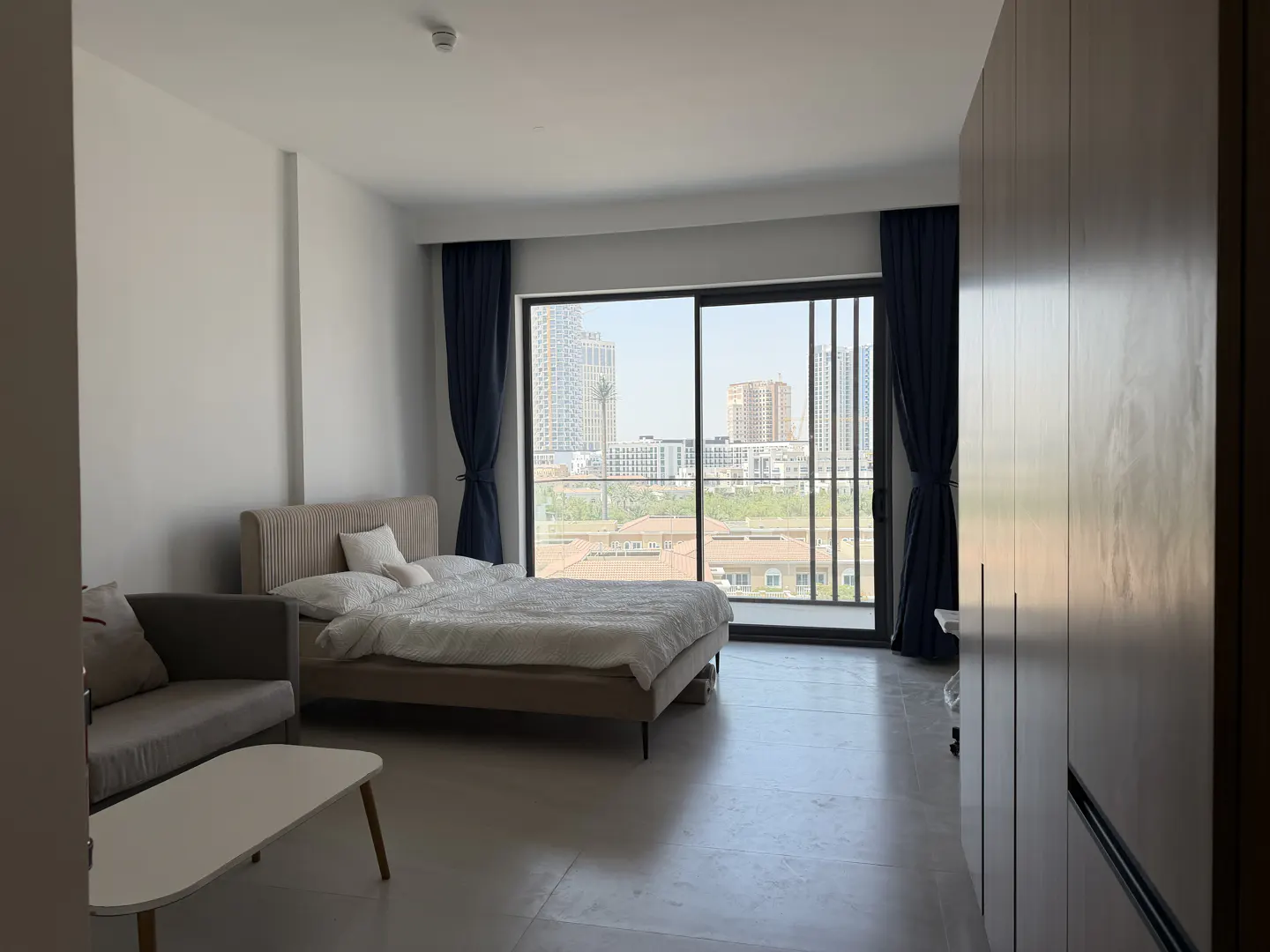 Modern bedroom with a beige bed, grey sofa, white coffee table, and large window overlooking a city skyline.