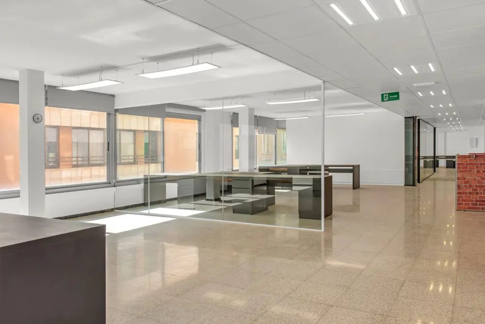 Bright, empty office space with desks, glass partitions, and large windows. Beige tile floor reflects light. An exit sign is visible on the ceiling.