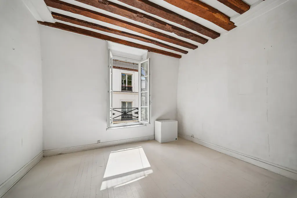 Bright, empty room with white walls, painted wood floor, and exposed wood beams. Open window shows a building across the street.