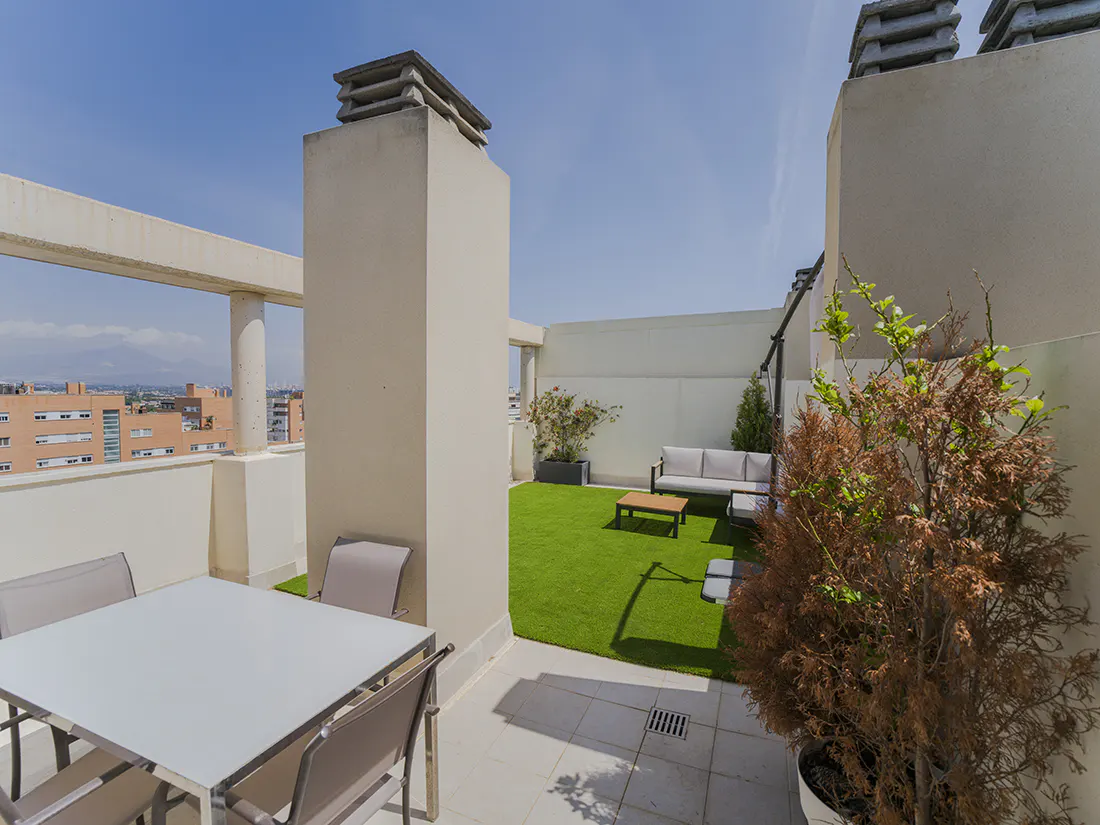 A rooftop patio with a table, chairs, and a seating area with a couch and coffee table. There is artificial grass and potted plants.