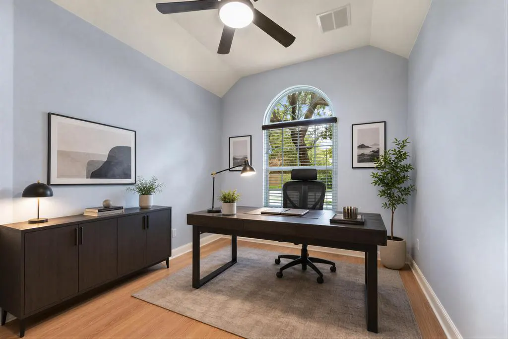 A home office with light blue walls, a dark desk, a black chair, and a large arched window.