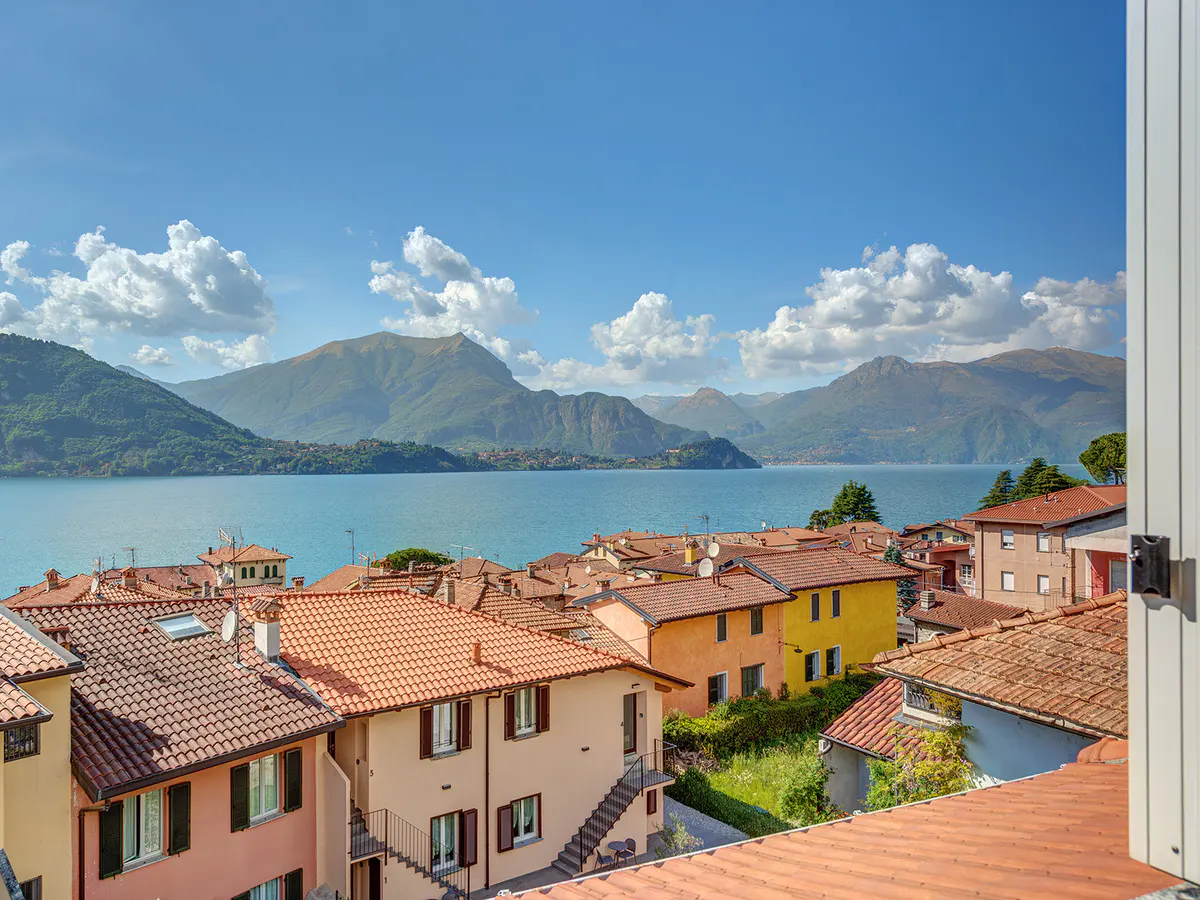 View from a window of colorful houses with red tile roofs overlooking a blue lake and mountains under a blue sky.