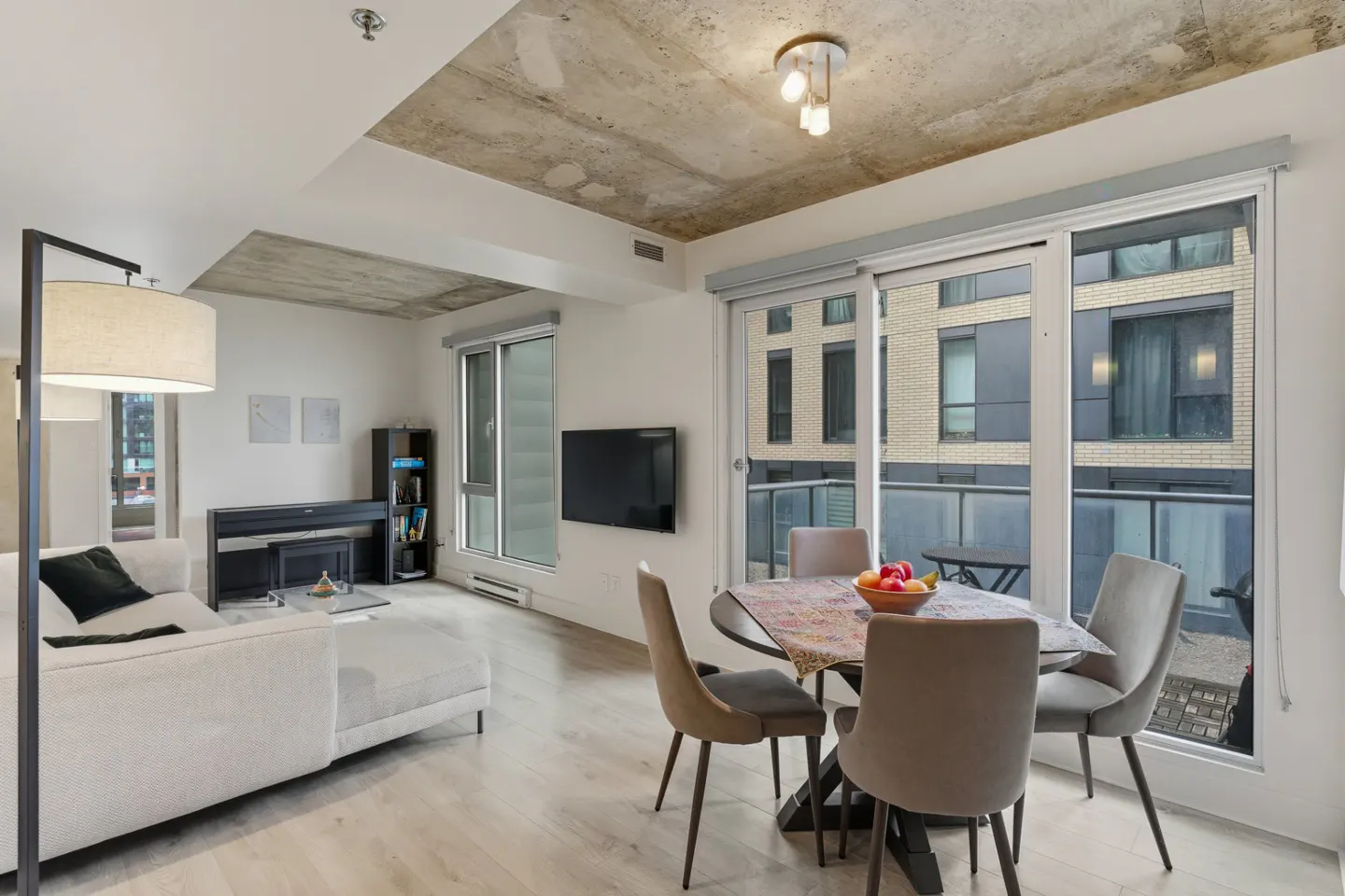 Bright, modern apartment with a white sofa, piano, and round dining table with four chairs. Large windows offer city views. Concrete ceiling.
