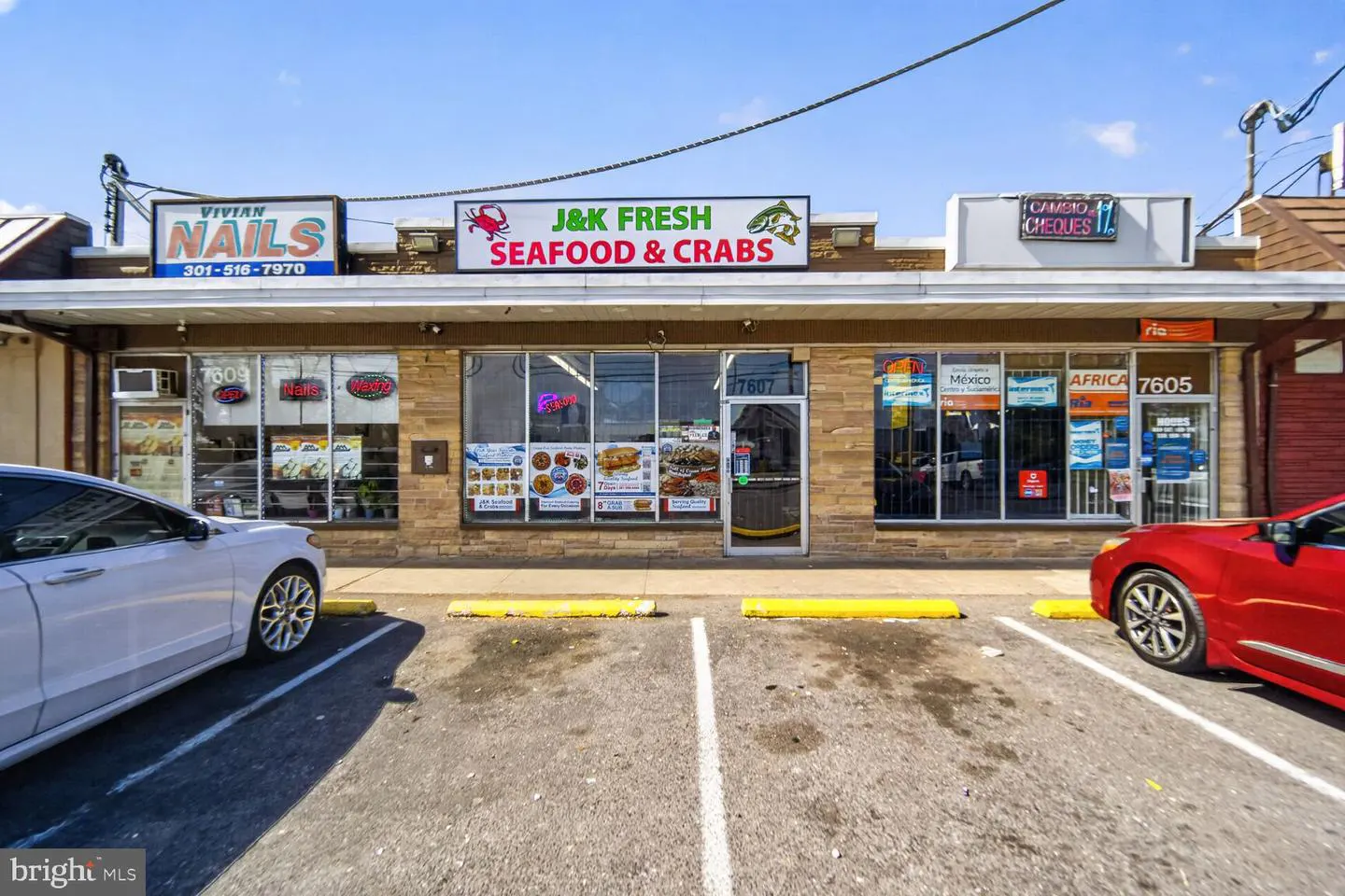 A one-story commercial building with a parking lot, featuring businesses like "J&K Fresh Seafood & Crabs" and "Vivian Nails." A white car is parked on the left, and a red car is on the right.