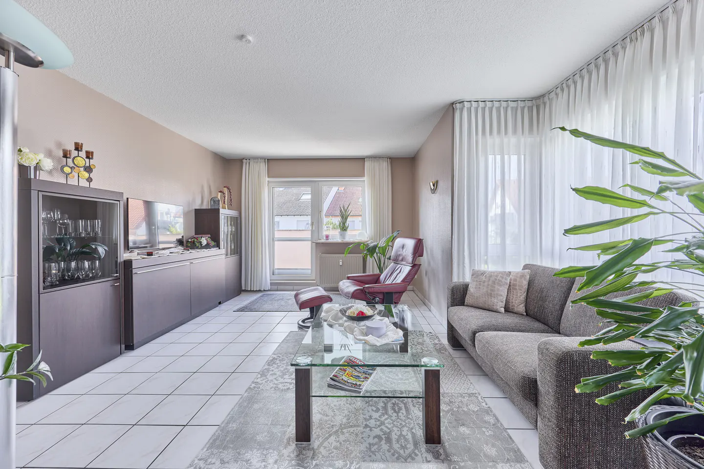 Living room with brown sofa, glass table, red recliner, and dark wood cabinets against a light pink wall. White tile floor and sheer curtains.