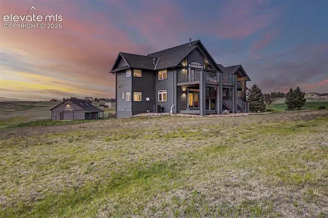 Dark gray multi-story home with a deck and detached garage in a grassy field under a colorful sunset sky.