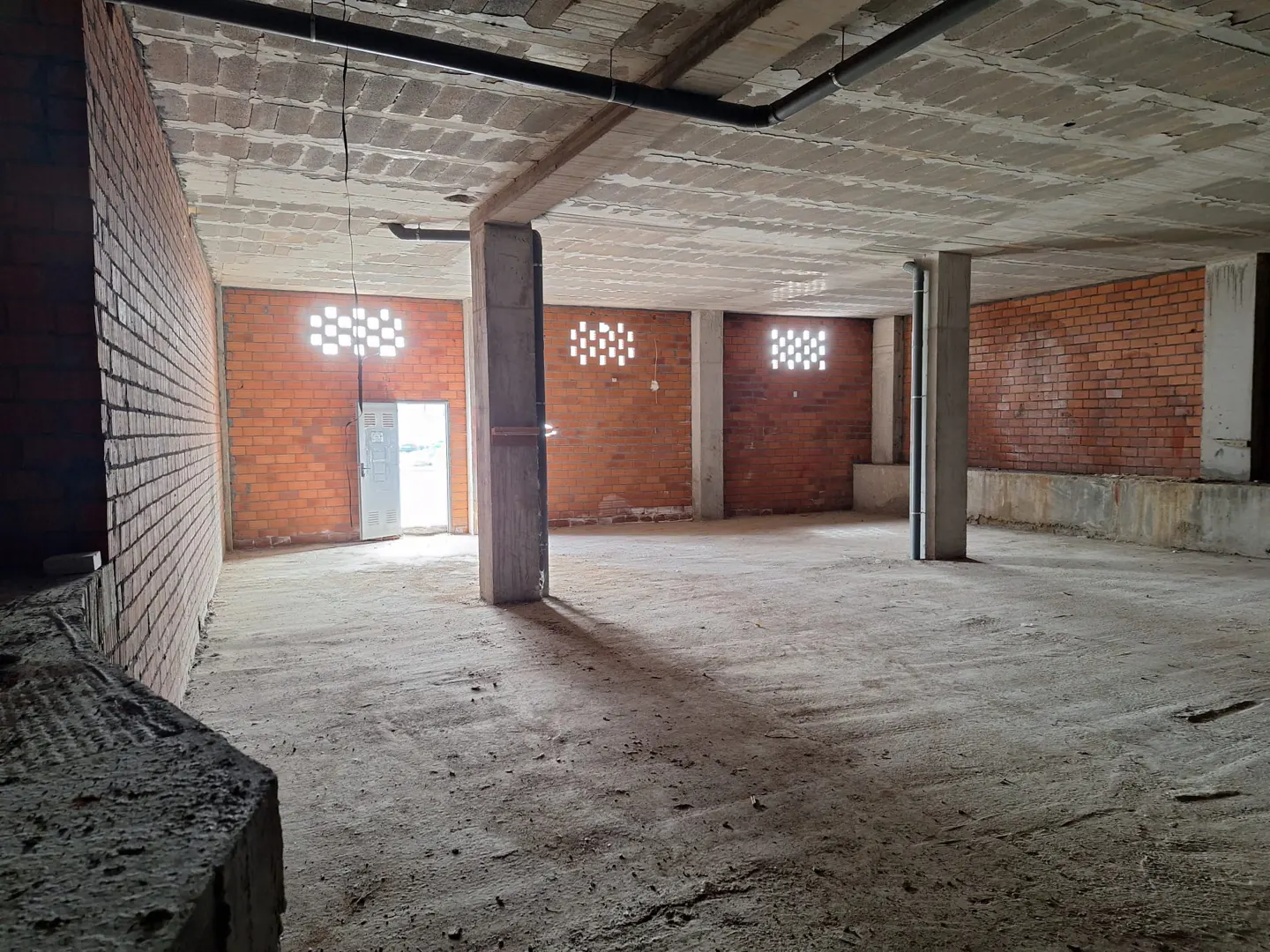 An unfinished, open-plan commercial space with exposed red brick walls, concrete pillars, and a rough concrete floor.