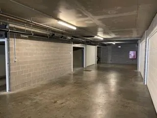 Indoor parking garage with concrete walls, floor, and ceiling. Fluorescent lights illuminate the space. Storage units line the walls.