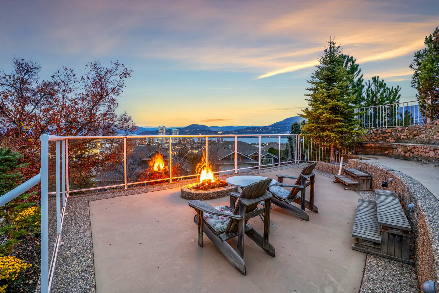Outdoor patio with a fire pit, chairs, and benches overlooking a city and lake at sunset.